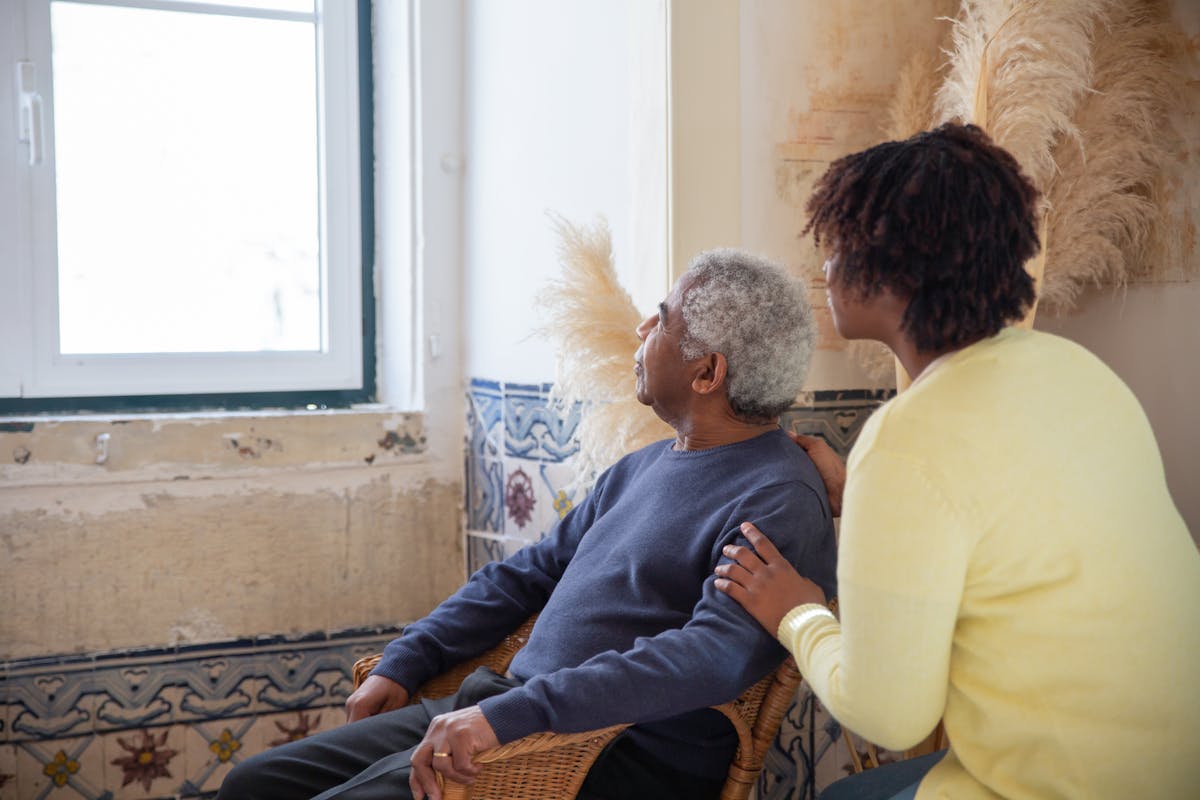 Older adult seated by a window with a caregiver beside them
